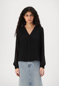 Svart bluse med V-hals, med gjennomsiktige lange ermer prydet med sm�å, teksturerte mønstre, kombinert med en lyseblå denimskjørt.