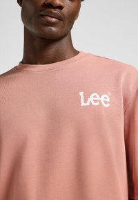 Lichtroze sweatshirt met lange mouwen, gemaakt van zachte stof, met een witte "Lee"-logo op de borst. Ronde halslijn en gestructureerde afwerking.