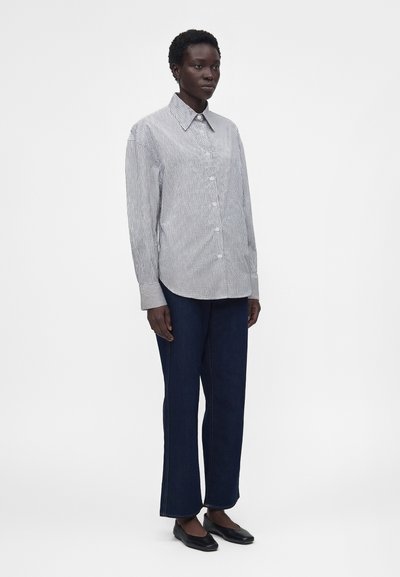 Γυναίκα φοράει ανοιχτό γκρι πουκάμισο με ρίγες και κουμπιά, σκούρο φαρδύ παντελόνι και μαύρα φλατ παπούτσια, στέκεται μπροστά σε απλό λευκό φόντο.