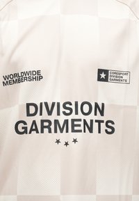Lys beige beklædning med rutenetmønster, prydet med sort tekst: "WORLDWIDE MEMBERSHIP" og "DIVISION GARMENTS." Inkluderer logoer og stjerner.
