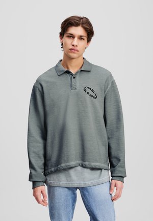 Gray, long-sleeve polo shirt with a collared neckline and embroidered logo. Worn over a lighter gray base layer, paired with light blue jeans.