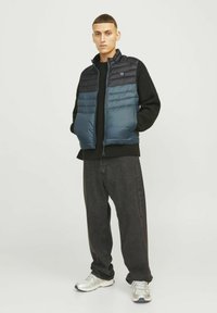 Vesta puffer albastră și neagră, cu un design matlasat orizontal și guler înalt, purtată peste un pulover negru texturat și asociată cu blugi largi de culoare închisă.