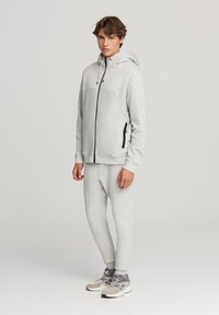 Giovane uomo in piedi che indossa una felpa con zip grigia chiara con dettagli neri, pantaloni tuta coordinati e sneakers grigie e beige.