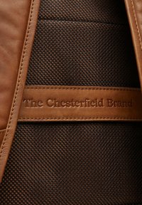Étiquette en cuir marron avec "The Chesterfield Brand" embossé, sur fond en maille texturée sombre. Design simple et fonctionnel.
