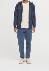 Homme portant un sweat à capuche bleu zippé, un t-shirt crème, un jean bleu et des baskets blanches avec des rayures brunes, debout devant un fond blanc.