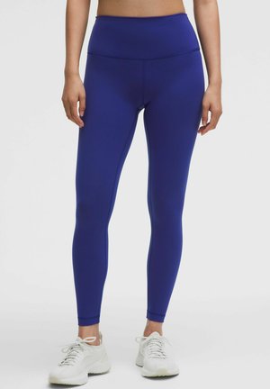 Woman wearing high-waisted, full-length royal blue leggings and white athletic shoes, standing against a plain light background.