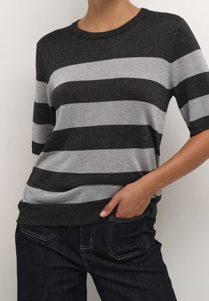 Personne portant un t-shirt à manches courtes noir et gris à rayures horizontales et un jean foncé, une main dans la poche, sur un fond uni.