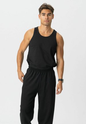 Mann stående vendt fremover iført en svart tanktop, svarte løse bukser og en armbåndsur mot en enkel lys bakgrunn.