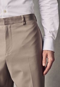 Beżowe, dopasowane spodnie o gładkiej fakturze, z płaskim przodem, bocznymi kieszeniami i paskiem w talii. Noszone z białą koszulą wizytową.