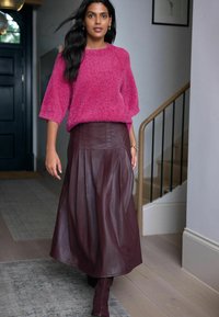 Flauschiger rosa Pullover mit weiten Ärmeln, kombiniert mit einem langen, bordeauxfarbenen Lederrock mit Falten. Modell trägt passende bordeauxfarbene knielange Stiefel.