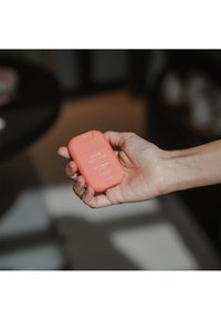 Contenitore di spray per bocca color corallo con angoli arrotondati, etichettato "HAAN MOUTH SPRAY Life's a Beach", tenuto in una mano contro uno sfondo sfocato.