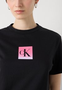 Melnā t-kreklā ir kvadrātveida logotips ar gradientu no rozā līdz baltai, kas rāda "cK" treknajās melnās burtiņās. Īsas piedurknes un apaļa kakla izgriezums.