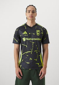 Camiseta deportiva negra con un llamativo patrón de salpicaduras en amarillo neón, con el logo de Adidas, emblema del Columbus Crew y un diseño de cuello en V.