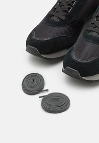 Chaussures de sport noires avec un mélange de daim et de maille, dotées de lacets plats gris foncé et d'une semelle en caoutchouc texturée.