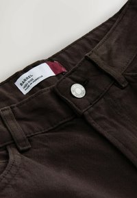 Tumši brūnas augsta vidukļa bikses ar redzamu pogu, uz kuras rakstīts "NEXT DENIM SINCE 1982", un iekšējo etiķeti ar uzrakstu "BARREL HIGH RISE MADE IN MOROCCO".