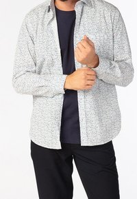 Koszula w kwiatowy wzór z lekkiego materiału, biała baza z niebieskimi akcentami, długie rękawy, zapinana na guziki, noszona na granatowej bluzie.