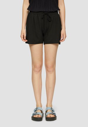 Femme portant un short noir à cordon, un haut noir et des sandales à double bride bleu clair, debout devant un fond uni gris clair.