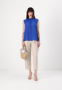 Blauwe mouwloze blouse met een gerimpelde halslijn, gecombineerd met lichtbeige broek. De persoon houdt een geweven rieten tas vast met een klein label.