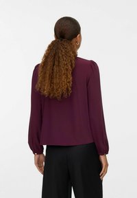 Blusa bordeaux con maniche lunghe a sbuffo e vestibilità morbida; dettaglio dei bottoni sulle maniche, texture liscia e scollatura rotonda.