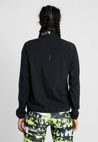 Nainen, jolla on letitetyt hiukset, pukeutuneena mustaan juoksutakkiin ja neonvihreisiin, mustiin ja valkoisiin kuvioituihin legginsseihin, nähtynä takaa.