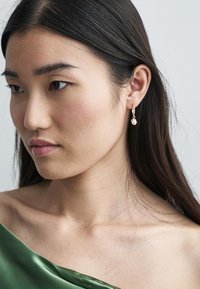 Χρυσό σκουλαρίκι με πέρλα, φορεμένο από άτομο με μακριά σκούρα μαλλιά, σε ουδέτερο φόντο. Λείο δέρμα και πράσινο ύφασμα.