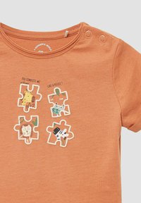Rdzawoczerwony t-shirt z czterema graficznymi zwierzętami w kształcie puzzli. Wykonany z miękkiego materiału z napami na ramieniu, co ułatwia zakładanie.