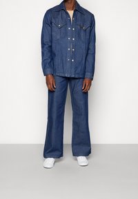 Blå denimjacka med spetsig krage, tryckknappar och två bröstfickor, i kombination med vida blå denimbyxor och vita sneakers.