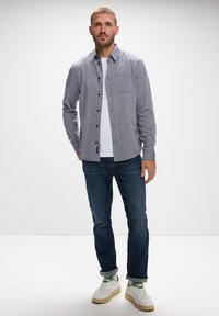 Camisa de botón gris con estampado y puntos blancos, combinada con una camiseta blanca, jeans oscuros y zapatillas blancas. Detalle de bolsillo frontal sencillo.