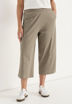 Person iført beige vidde-cropped bukser med lommer og hvide sneakers med brune striber, stående mod en lys baggrund.