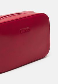 Red faux leather rectangular bag with a zipper closure and embossed logo. Smooth texture with rounded edges and visible stitching.