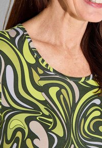 Haut vert avec un motif abstrait vibrant comportant des tourbillons en citron vert, vert foncé et beige clair, avec un col rond et une texture douce.