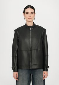 Jachetă din piele neagră cu guler înalt, închidere cu fermoar argintiu și accente cu cataramă pe mâneci. Textură netedă și design structurat.