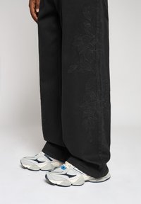 Pantalon en jean noir avec broderie florale sur le côté, associé à des baskets grises et bleues au design massif et à la surface texturée.