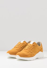 Yellow suede sneakers with mesh panels, white rubber soles, and round laces. Features a logo on the tongue and side stitching detail.