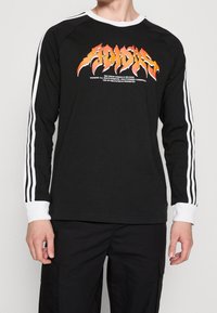 Svart långärmad skjorta med vit krage och manschetter, prydd med grafiska text i orange låga och vita ränder som löper längs ärmarna.