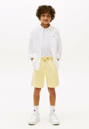Kurzärmliges weißes Hemd über einem weißen T-Shirt, kombiniert mit gelben Shorts, weißen Turnschuhen und gemusterten Socken.