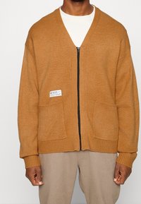 Brun stickad kofta med dragkedjefram, två framfickor, ribbstickade muddar och fåll. Har en etikett på vänstra fickan.