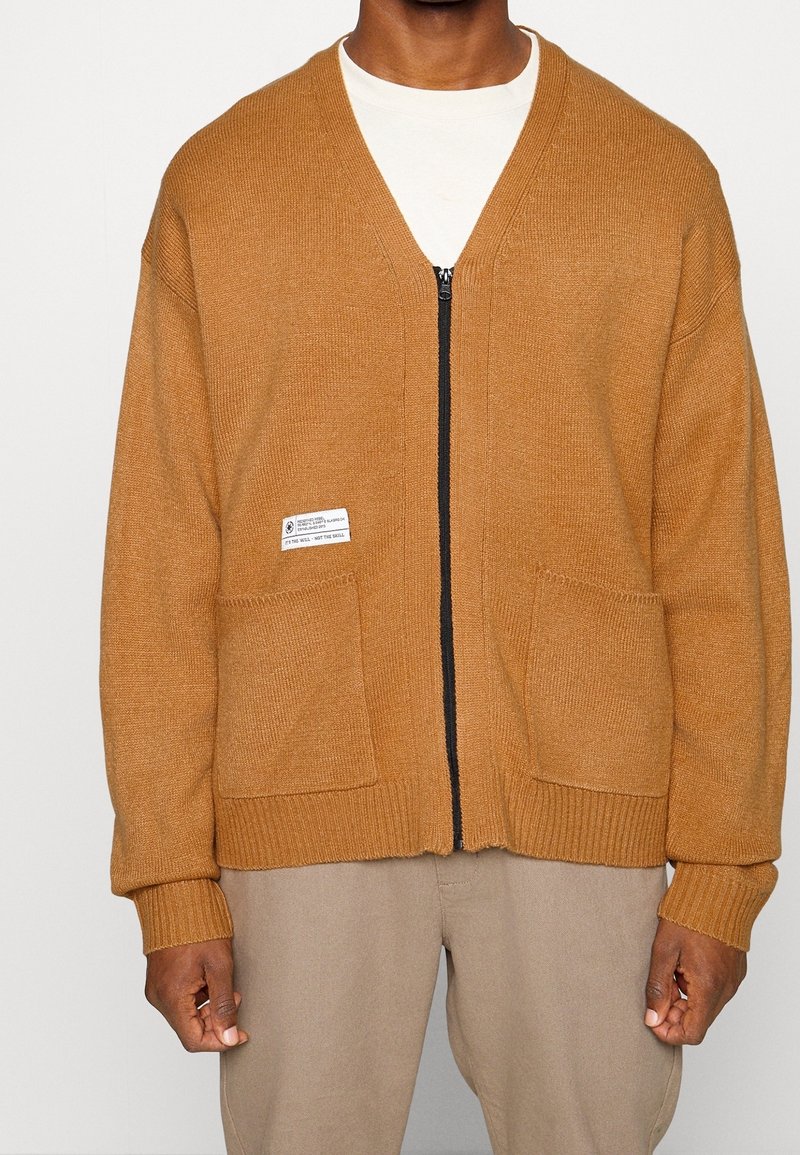 Brun stickad kofta med dragkedjefram, två framfickor, ribbstickade muddar och fåll. Har en etikett på vänstra fickan.