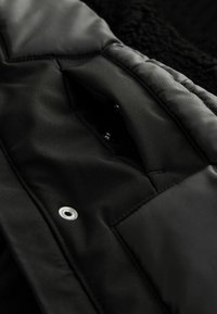 Schwarze gepolsterte Jackeninnenseite aus glattem Kunstfasermaterial, mit einer Reißverschlusstasche und einem metallischen Druckknopf. Weiche Textur im Kontrast zum anderen Material.