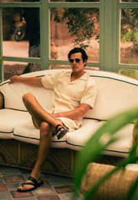 Rieten sofa met witte kussens voorzien van groene rand, bezet door een man in een crèmekleurig shirt en shorts, die een zonnebril en zwarte sandalen draagt.