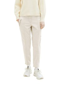 Personne portant un pantalon beige zippé, un pull crème, des chaussettes blanches et des baskets blanches, debout avec les mains détendues le long du corps.