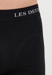 Crne bokserice s glatkom teksturom, s tamnim pojasom na kojem je bijelim bojama izvezeno "LES DEUX". Vidljivi su detalji sa strane.