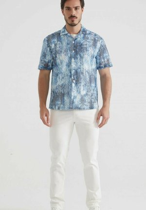 Chemise à manches courtes, à boutons, dans des teintes de bleu et blanc, avec un motif abstrait texturé. Associée à un pantalon blanc.