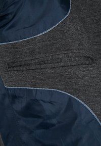 Tela texturizada gris con detalles en azul oscuro, presenta un bolsillo oculto y costuras en contraste a lo largo de los bordes. Diseño de ropa exterior casual.