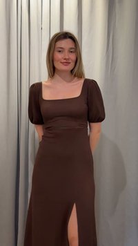 Kvinde med glat blondt hår iført en brun kjole med puffede ærmer og et frontslids, stående mod en lysefarvet baggrund med gardin.