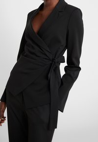 Svart omslag blazer tillverkad av slätt tyg, med djup V-ringning, långa ärmar och en knytlinje för justering av passform.