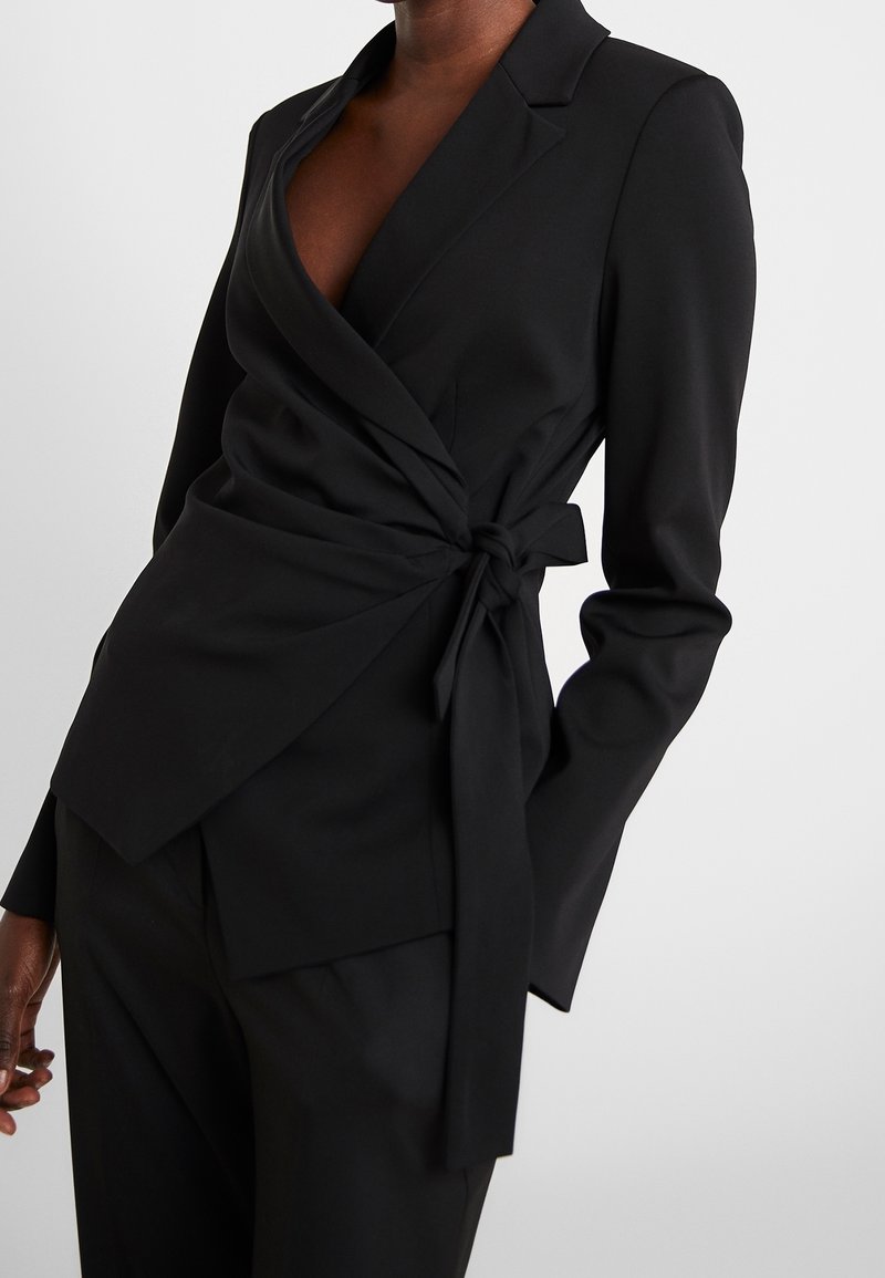 Svart omslag blazer tillverkad av slätt tyg, med djup V-ringning, långa ärmar och en knytlinje för justering av passform.