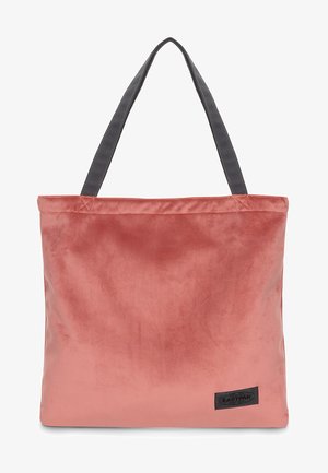 Roze fluwelen tote bag met grijze stoffen handvatten, vierkante vorm en een gestikt logo patch in de rechteronderhoek. Soepele textuur.