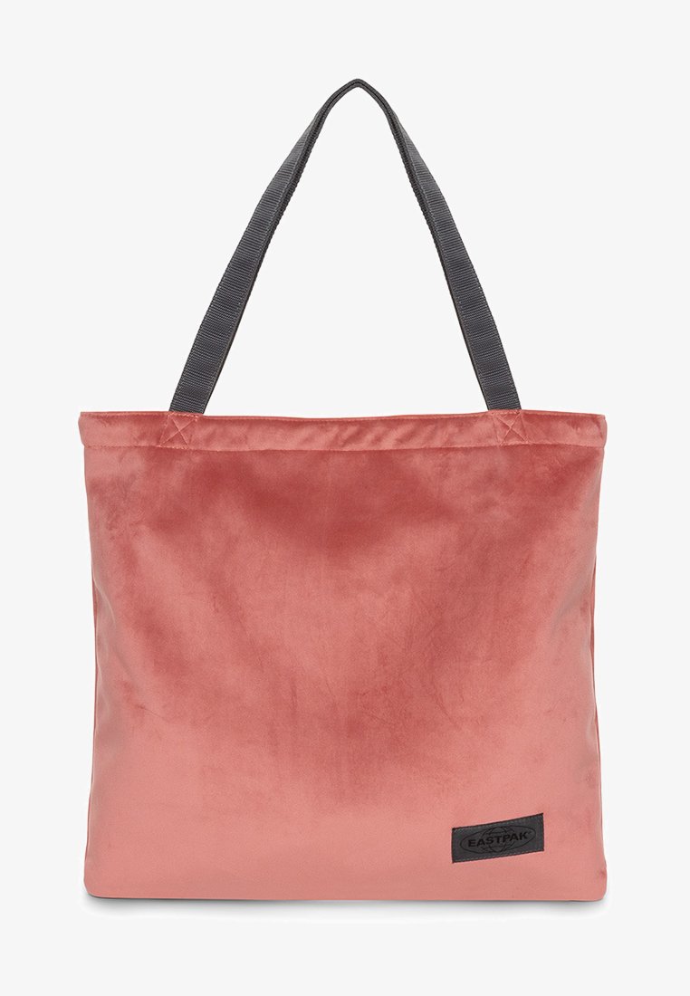 Pink velvet tote bag with gray fabric handles, square shape, and a stitched logo patch on the bottom right corner. Smooth texture.