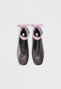 Pair of dark brown leather ballet flats with pink ankle ribbons and "Tommy Jeans" logo on toes, viewed from above on white background.